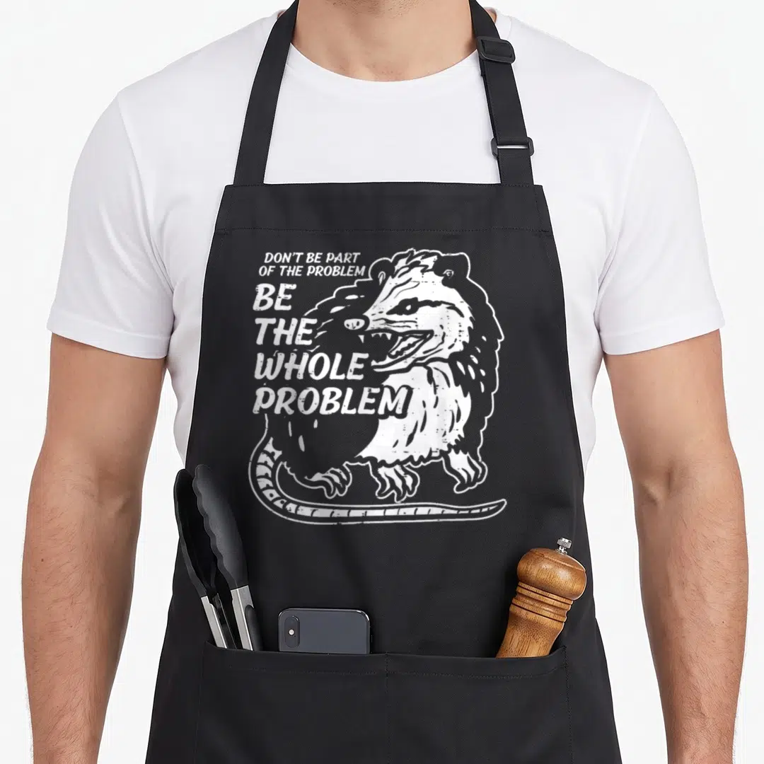 Opossum parts of an apron - White distressed raccoon illustration with stacked text Don't Be Part Of The Problem Be — shown on a black cotton canvas apron against a white background