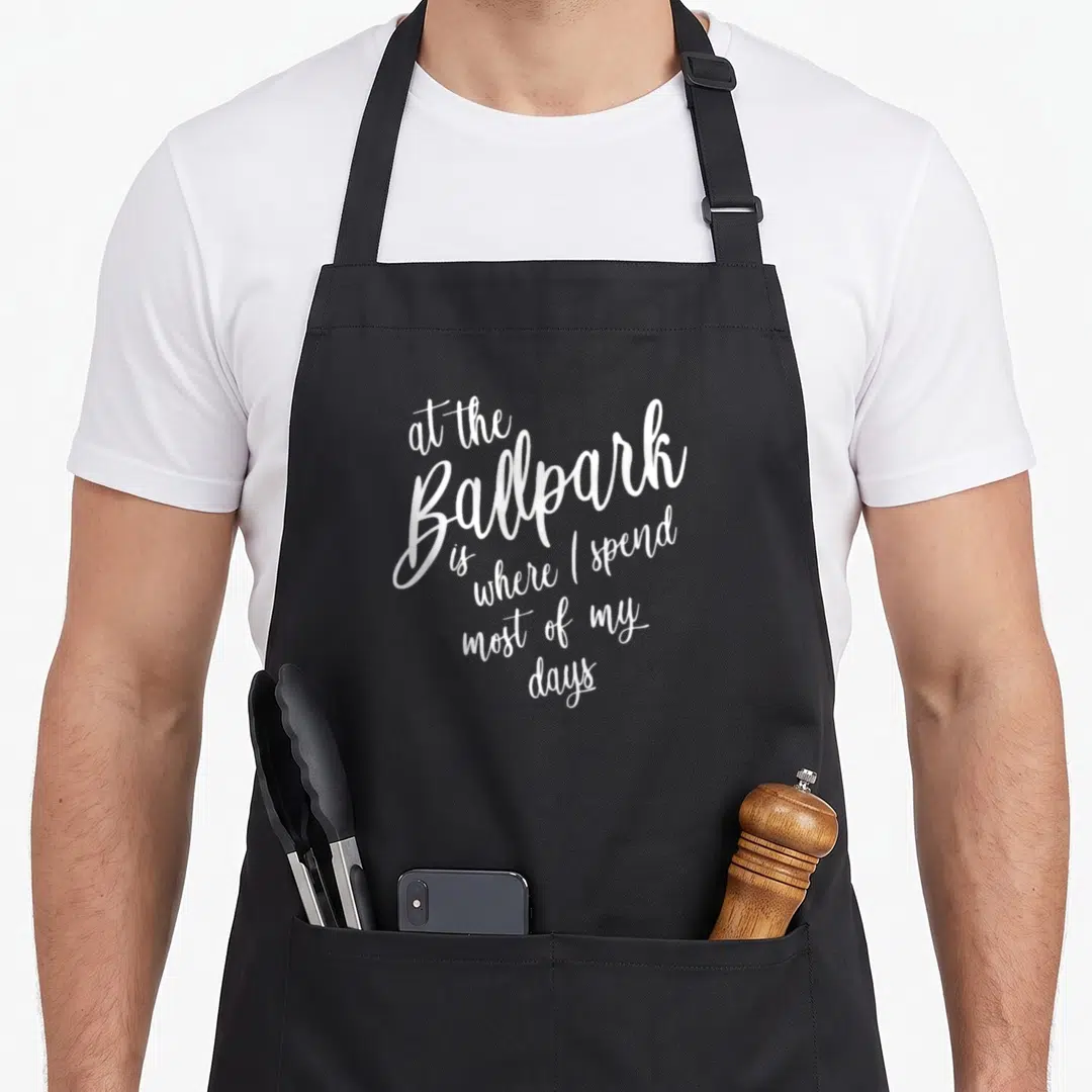 Spend baseball apron - White stacked script text reading at the Ballpark is where I spend most of my days — shown on a black cotton canvas apron against a white background
