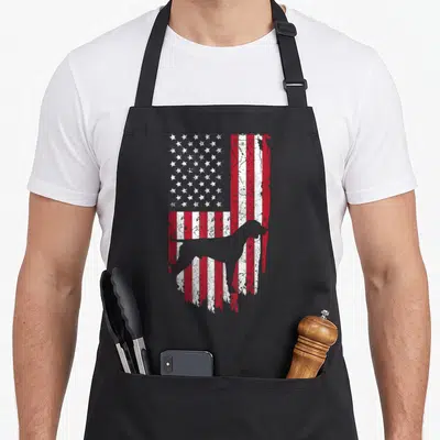 German haired pointer apron - Grey dog silhouette standing on a distressed red white and blue American flag — shown on a black cotton canvas apron against a white background