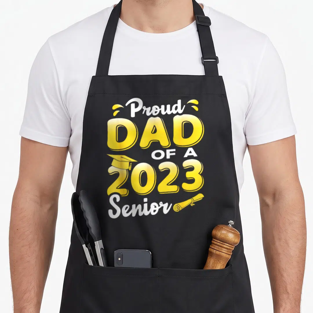 Proud funny graduation apron - Stacked yellow and white text reading Proud DAD OF A 2023 Senior with cap and diploma — shown on a black cotton canvas apron against a white background
