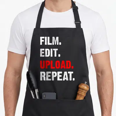 Film video editing apron - Stacked white and red text reading FILM EDIT UPLOAD REPEAT in bold distressed font — shown on a black cotton canvas apron against a white background