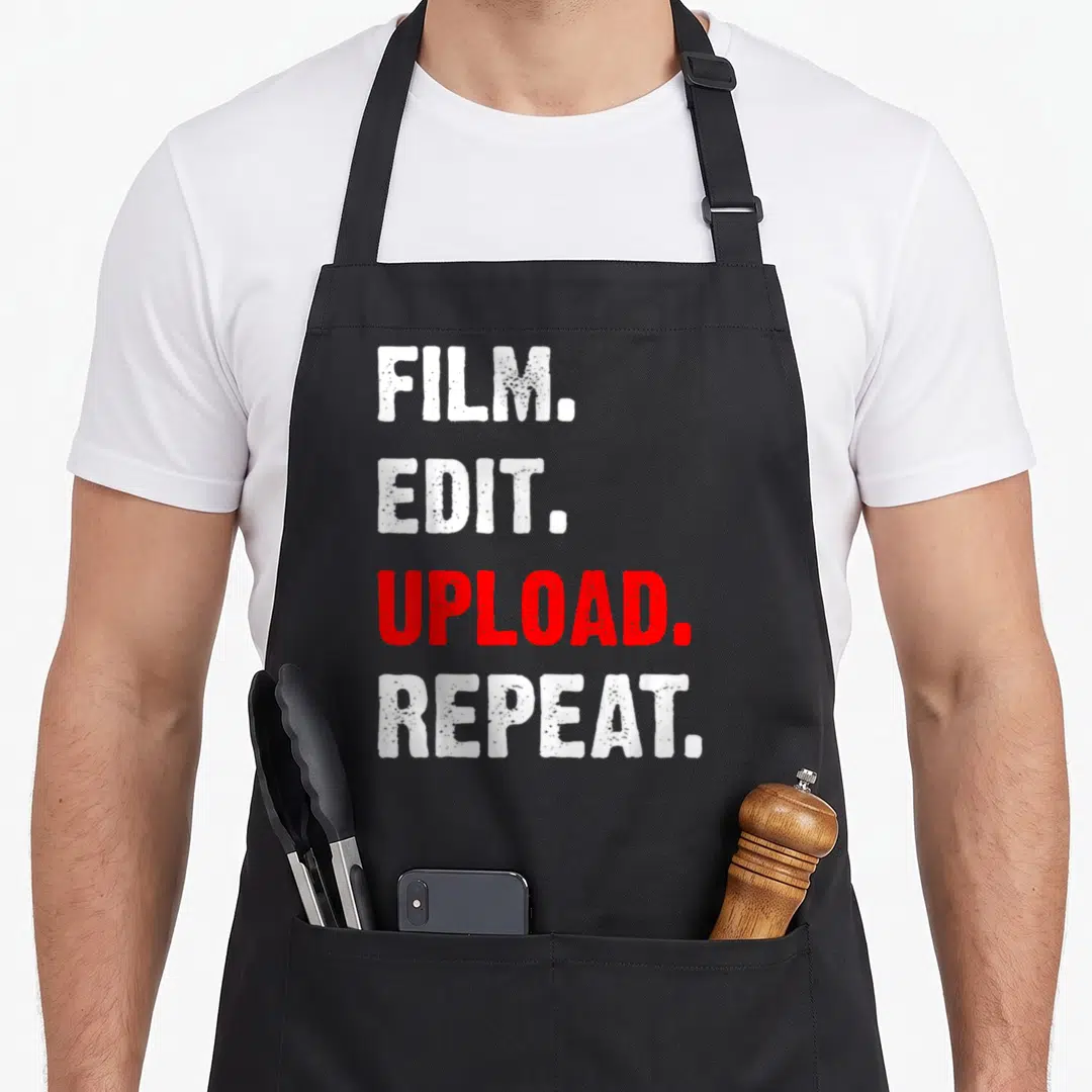 Film video editing apron - Stacked white and red text reading FILM EDIT UPLOAD REPEAT in bold distressed font — shown on a black cotton canvas apron against a white background