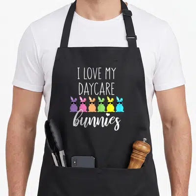 Easter daycare teacher aprons - Six colorful cartoon bunnies wearing sunglasses below stacked white text reading I Love — shown on a black cotton canvas apron against a white background