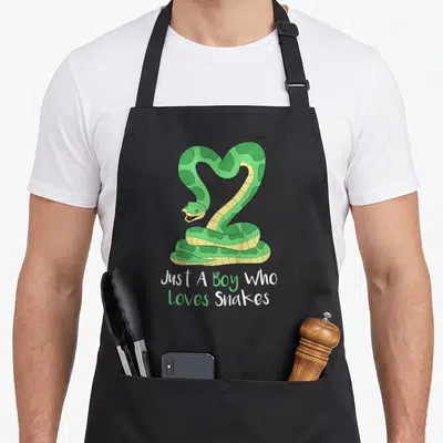 Snake boa apron - Cartoon green snake coiled above white and green text reading Just A Boy Who Loves Snakes — shown on a black cotton canvas apron against a white background
