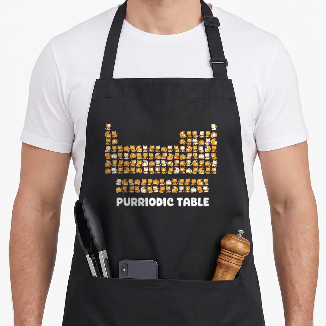 Table cat chemistry apron - Cluster of orange and white cartoon cats arranged in a periodic table grid above white text — shown on a black cotton canvas apron against a white background