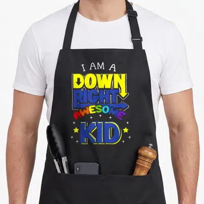 Trisonomy chromosomes apron - Stacked colorful text I AM A DOWN RIGHT AWESOME KID with arrows and scattered stars — shown on a black cotton canvas apron against a white background