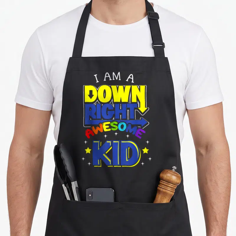 Trisonomy chromosomes apron - Stacked colorful text I AM A DOWN RIGHT AWESOME KID with arrows and scattered stars — shown on a black cotton canvas apron against a white background