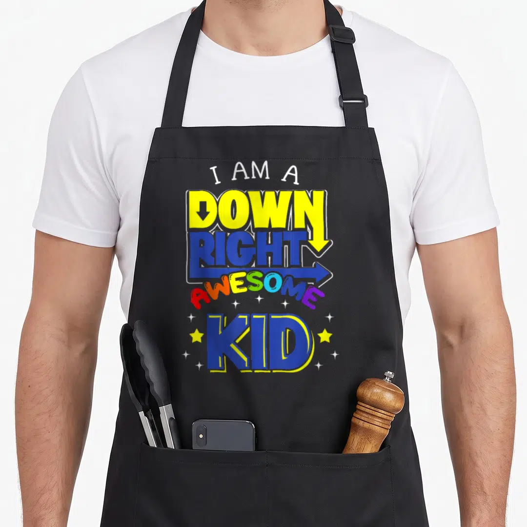 Trisonomy chromosomes apron - Stacked colorful text I AM A DOWN RIGHT AWESOME KID with arrows and scattered stars — shown on a black cotton canvas apron against a white background