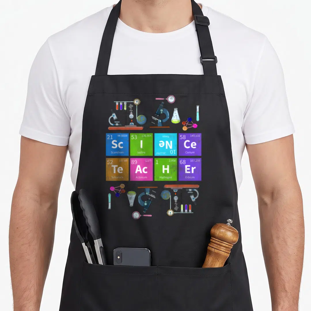 Teacher table apron - Cartoon microscopes and flasks frame a grid of eight colored periodic table tiles showing element — shown on a black cotton canvas apron against a white background