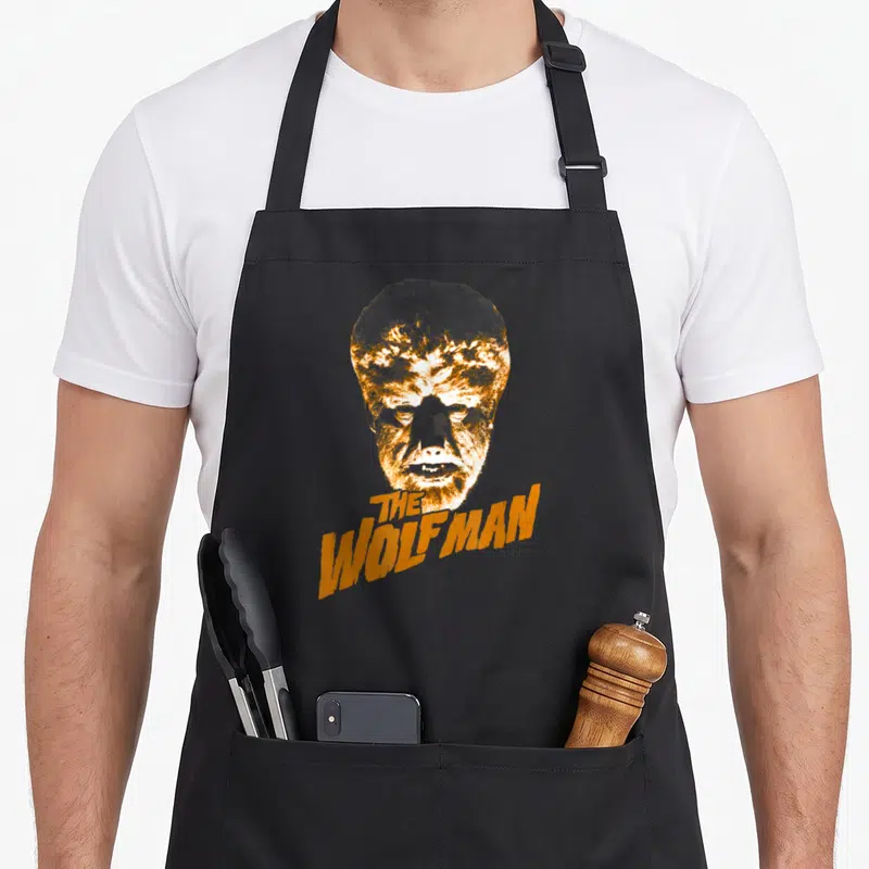 Wolf universal monsters apron - Distressed orange and white wolfman head above THE WOLFMAN text on black fabric — shown on a black cotton canvas apron against a white background