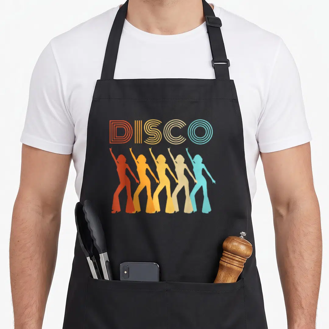 Disco diva 70s apron - Five female silhouettes in disco poses under striped DISCO text in orange yellow beige and light — shown on a black cotton canvas apron against a white background