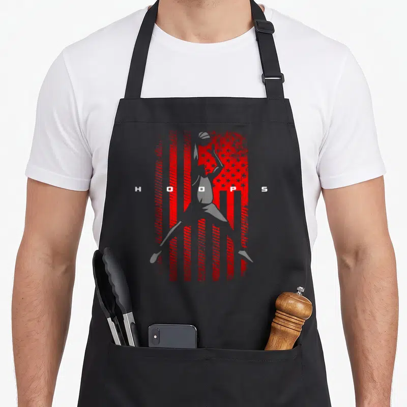 Cute basketball boys apron - White basketball player silhouette over distressed red white flag with HOOPS text — shown on a black cotton canvas apron against a white background