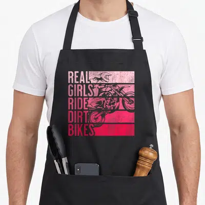 Ride dirt bike apron - stacked pink and red text reading real girls ride dirt bikes next to a distressed rider — shown on a black cotton canvas apron against a white background