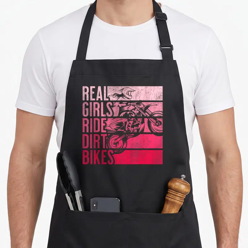 Ride dirt bike apron - stacked pink and red text reading real girls ride dirt bikes next to a distressed rider — shown on a black cotton canvas apron against a white background