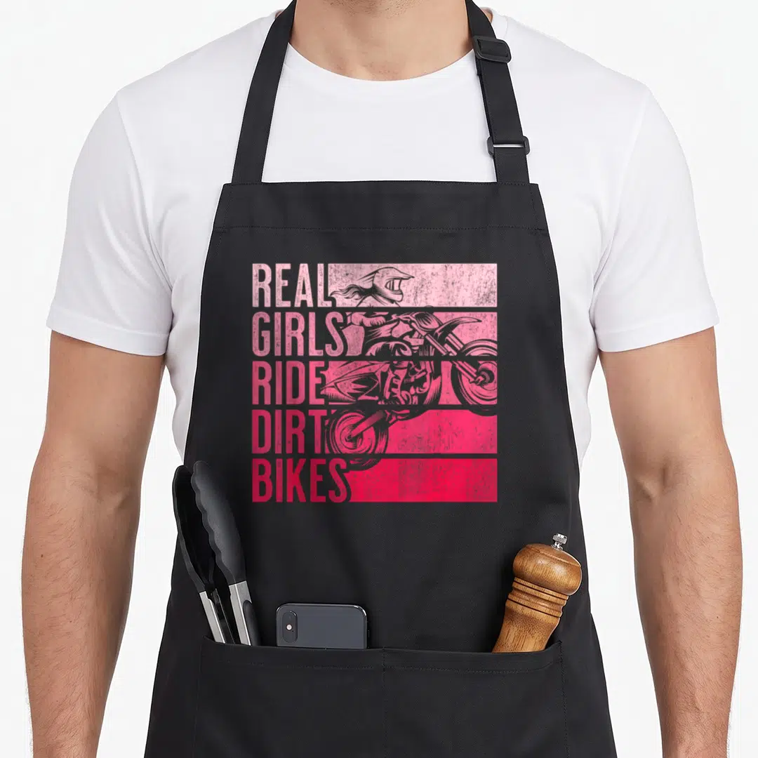 Ride dirt bike apron - stacked pink and red text reading real girls ride dirt bikes next to a distressed rider — shown on a black cotton canvas apron against a white background