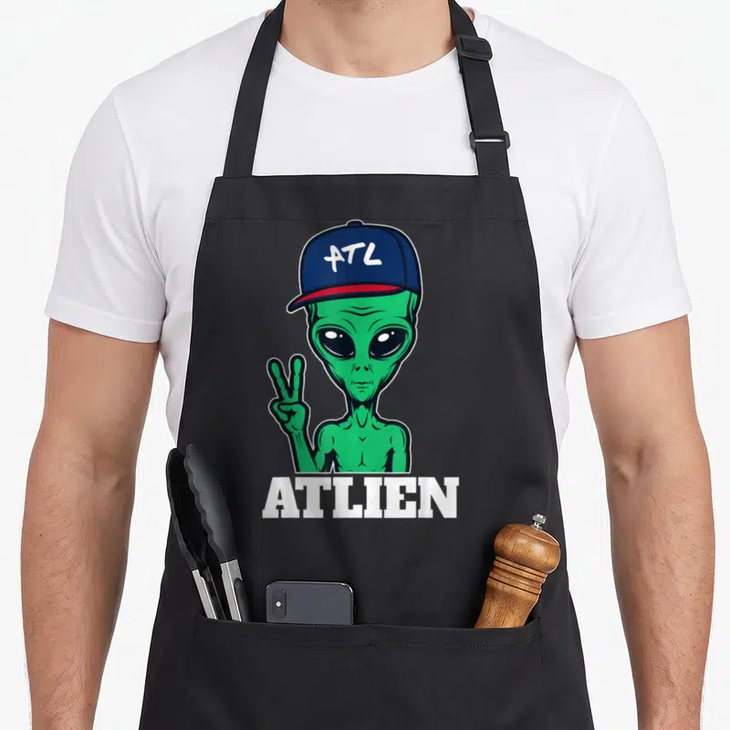 Atl atlanta apron - Green cartoon alien in blue cap with ATL text and ATLIEN word below — shown on a black cotton canvas apron against a white background
