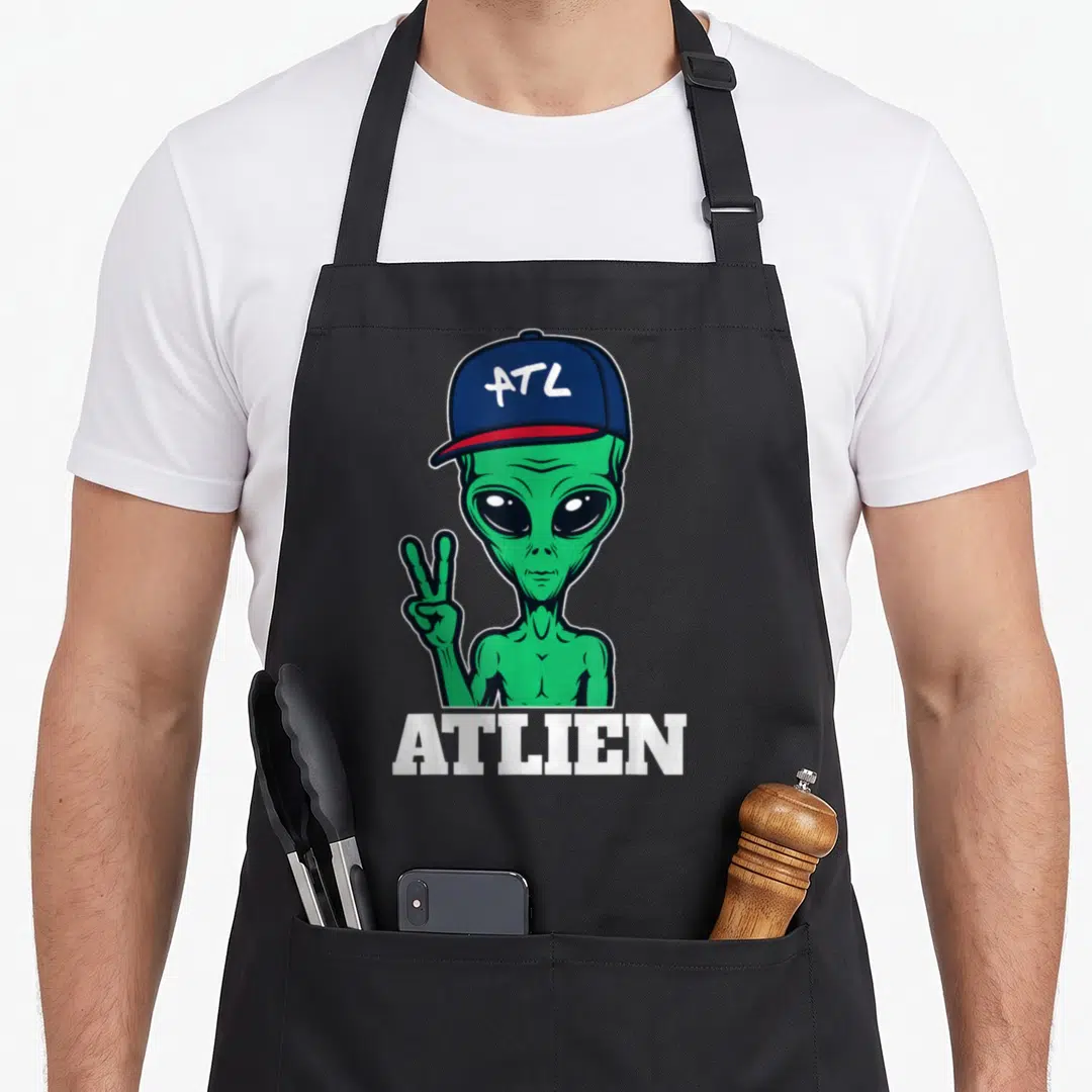 Atl atlanta apron - Green cartoon alien in blue cap with ATL text and ATLIEN word below — shown on a black cotton canvas apron against a white background