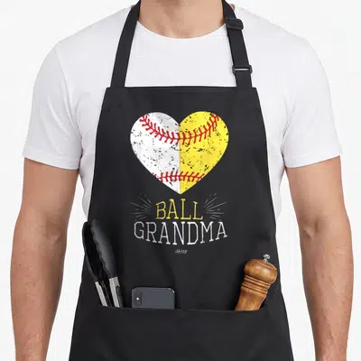 Ball grandma apron - Split white and yellow heart with red stitching above distressed BALL GRANDMA text — shown on a black cotton canvas apron against a white background