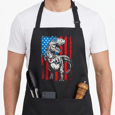Trex 4th of july apron - T-Rex wearing sunglasses holding a red white blue popsicle in front of a distressed flag — shown on a black cotton canvas apron against a white background