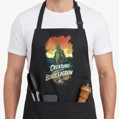Universal ogre apron - Green creature standing in water against orange sunset with white stacked text reading Creature — shown on a black cotton canvas apron against a white background
