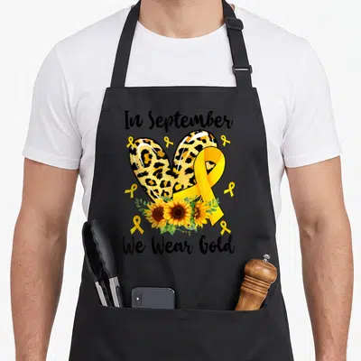 Funny september cancer awareness apron - Leopard heart, yellow ribbon, sunflowers, and text In September We Wear Gold — shown on a black cotton canvas apron against a white background