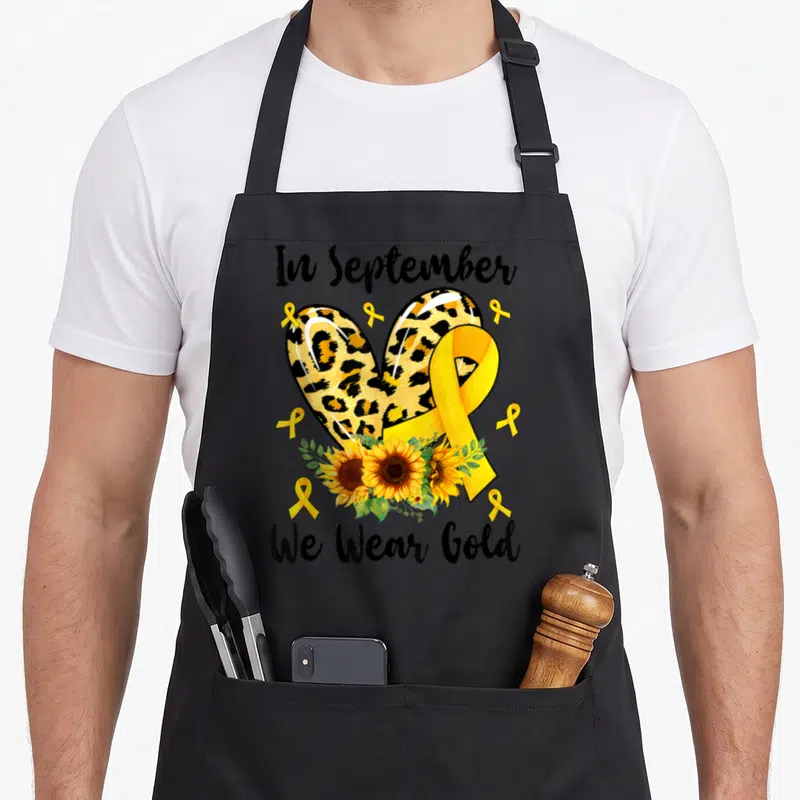 Funny september cancer awareness apron - Leopard heart, yellow ribbon, sunflowers, and text In September We Wear Gold — shown on a black cotton canvas apron against a white background