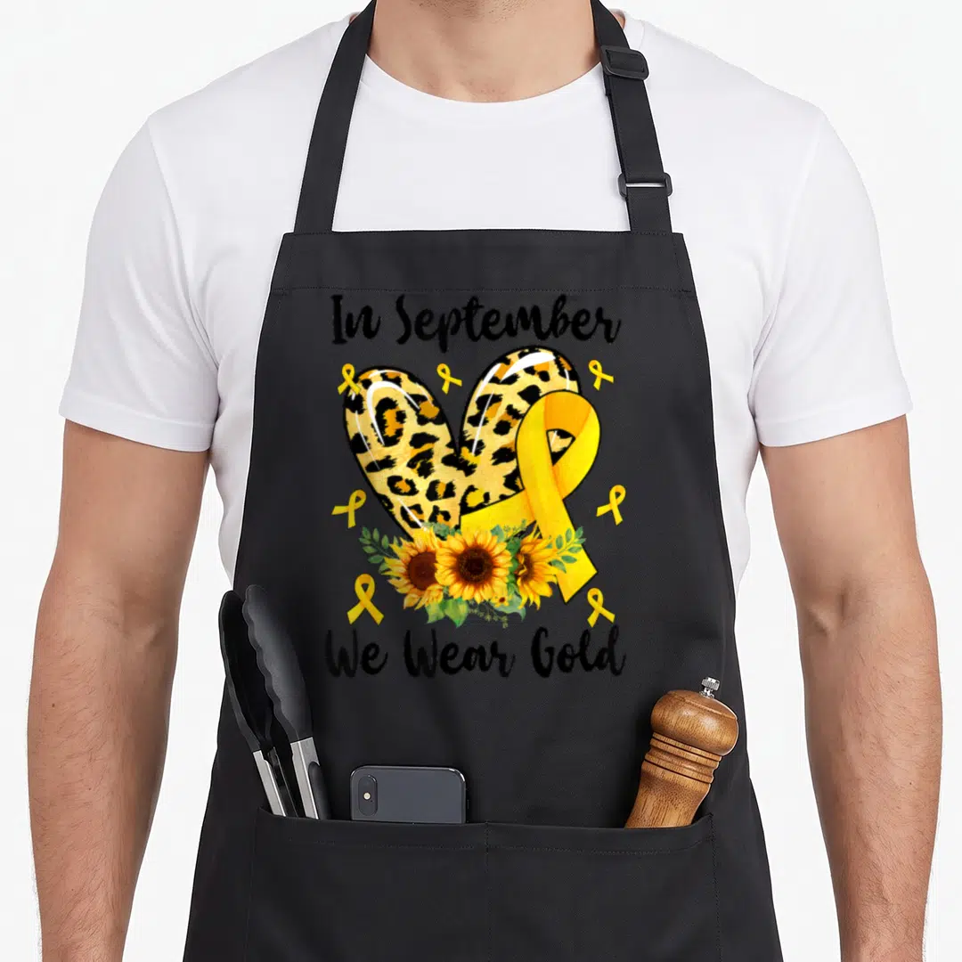 Funny september cancer awareness apron - Leopard heart, yellow ribbon, sunflowers, and text In September We Wear Gold — shown on a black cotton canvas apron against a white background