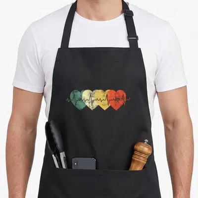 Nurse heartbeat nursing apron - Four overlapping hearts in green beige yellow and red with a running grey heartbeat line — shown on a black cotton canvas apron against a white background