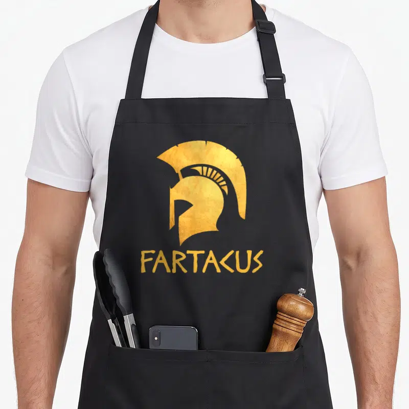 Fartacus joke apron - Gold minimalist Spartan helmet centered above the word FARTACUS in matching gold lettering — shown on a black cotton canvas apron against a white background