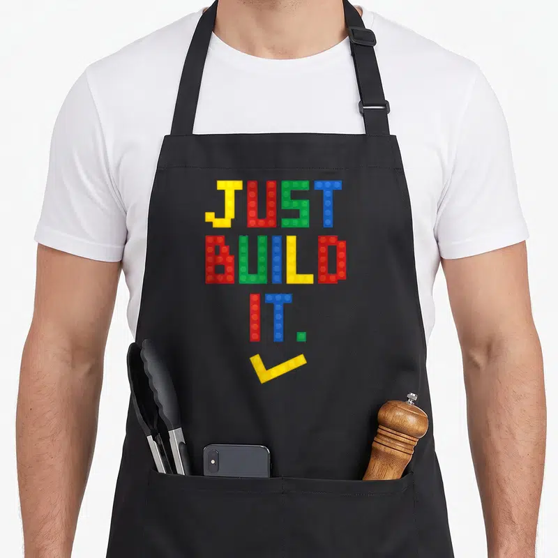 Build master building apron - Colorful building block text reading JUST BUILD IT. with a yellow checkmark below — shown on a black cotton canvas apron against a white background