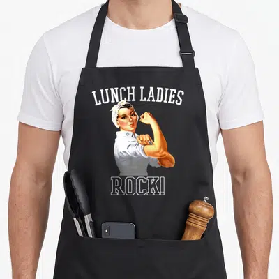 Rock cafeteria aprons - Retro woman flexing arm between LUNCH LADIES and ROCK! text in white tan yellow — shown on a black cotton canvas apron against a white background