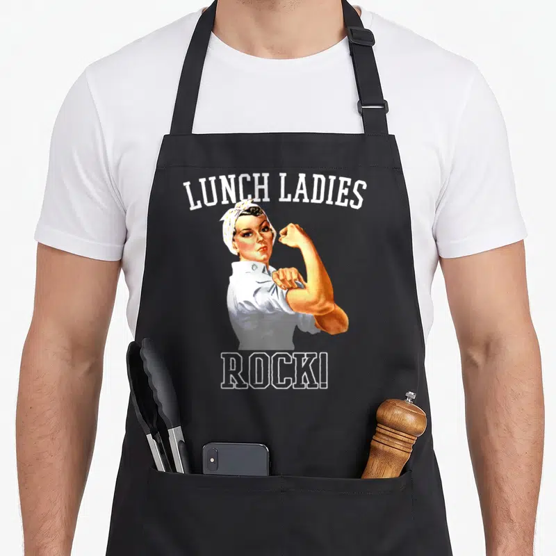 Rock cafeteria aprons - Retro woman flexing arm between LUNCH LADIES and ROCK! text in white tan yellow — shown on a black cotton canvas apron against a white background