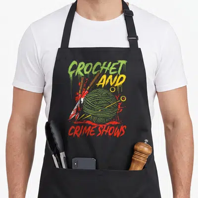Crime crochet apron - Green yarn ball pierced by hook and needles with red splatters and dripping text — shown on a black cotton canvas apron against a white background