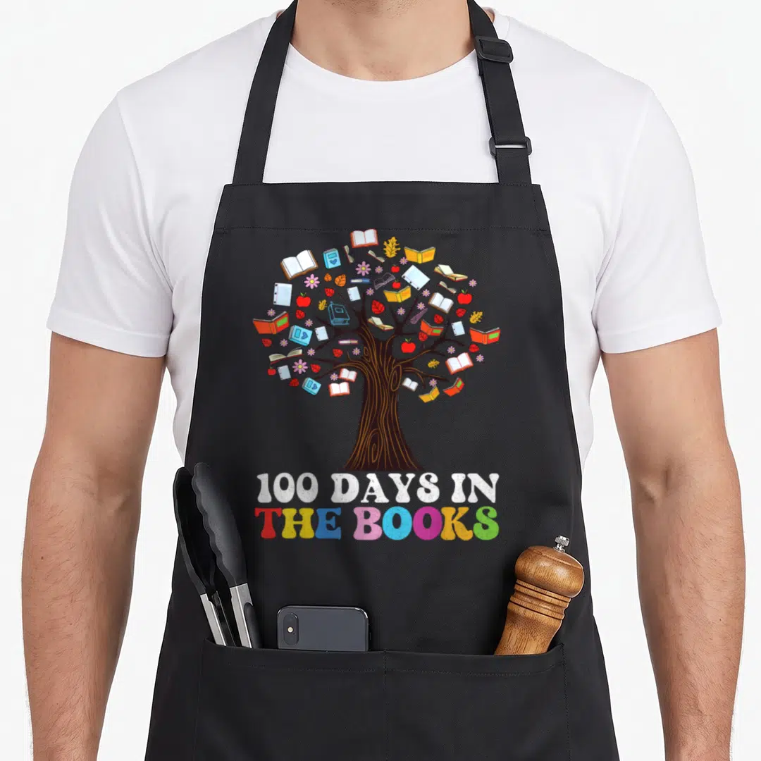 Book reading teacher apron - Cartoon tree with books apples and flowers above multi-colored 100 DAYS IN THE BOOKS text — shown on a black cotton canvas apron against a white background