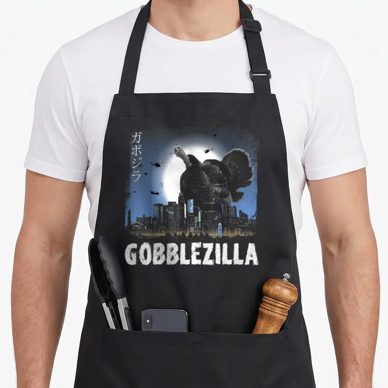 Gobblezilla thanksgiving day aprons - Giant turkey monster over city skyline with helicopters and white GOBBLEZILLA text — shown on a black cotton canvas apron against a white background