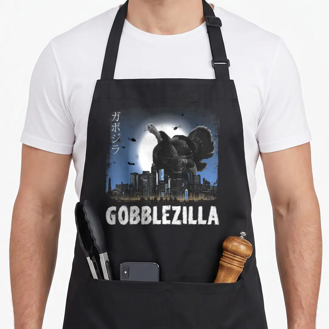 Gobblezilla thanksgiving day aprons - Giant turkey monster over city skyline with helicopters and white GOBBLEZILLA text — shown on a black cotton canvas apron against a white background