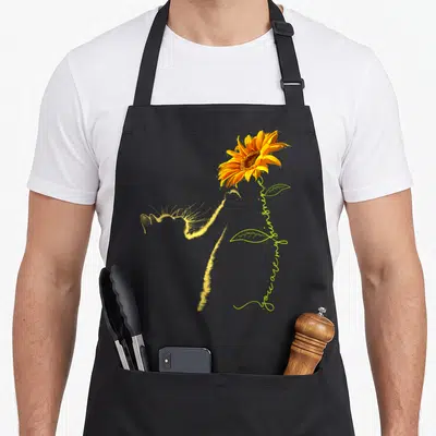 Sunflower cat lady apron - Yellow cat silhouette beside sunflower with you are my sunshine script — shown on a black cotton canvas apron against a white background