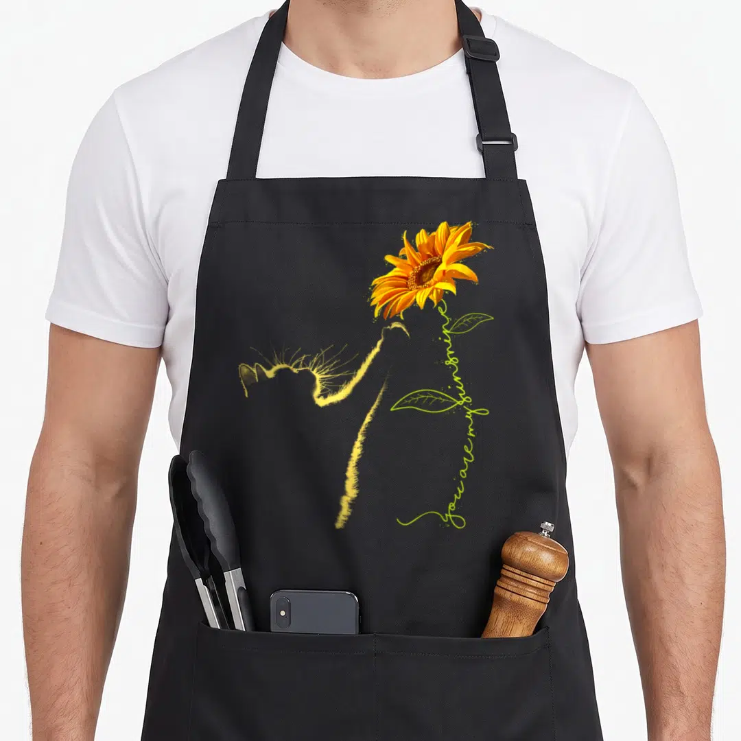 Sunflower cat lady apron - Yellow cat silhouette beside sunflower with you are my sunshine script — shown on a black cotton canvas apron against a white background