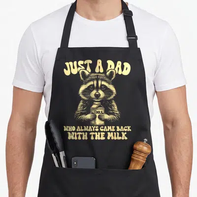 Funny raccoon milk aprons - Beige raccoon holding milk carton with arched Just A Dad text and stacked caption below — shown on a black cotton canvas apron against a white background