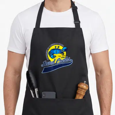 University crab apron - Blue crab holding yellow scales inside yellow circle with University of American Samoa Law Land — shown on a black cotton canvas apron against a white background