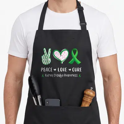 Peace kidney disease awareness apron - Green peace sign, pink heart, and ribbon with text PEACE LOVE CURE — shown on a black cotton canvas apron against a white background