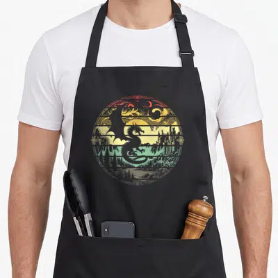 Playing master apron - Grey dragon silhouette inside circular frame with red yellow and green landscape bands — shown on a black cotton canvas apron against a white background