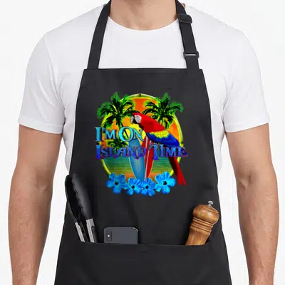 Island tropical print aprons - Cartoon parrot perched on surfboards inside a sunset circle with blue flowers and text — shown on a black cotton canvas apron against a white background