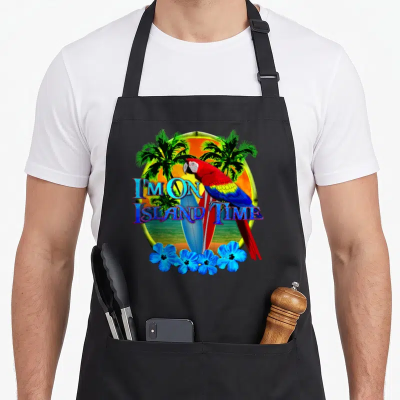 Island tropical print aprons - Cartoon parrot perched on surfboards inside a sunset circle with blue flowers and text — shown on a black cotton canvas apron against a white background