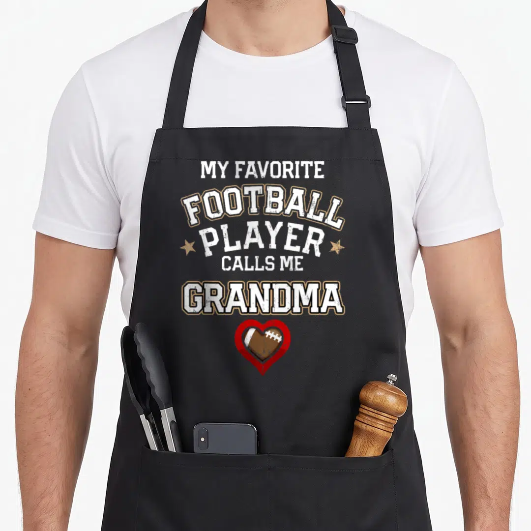 Player grandma aprons - Stacked text My Favorite Football Player Calls Me Grandma above red heart containing brown — shown on a black cotton canvas apron against a white background