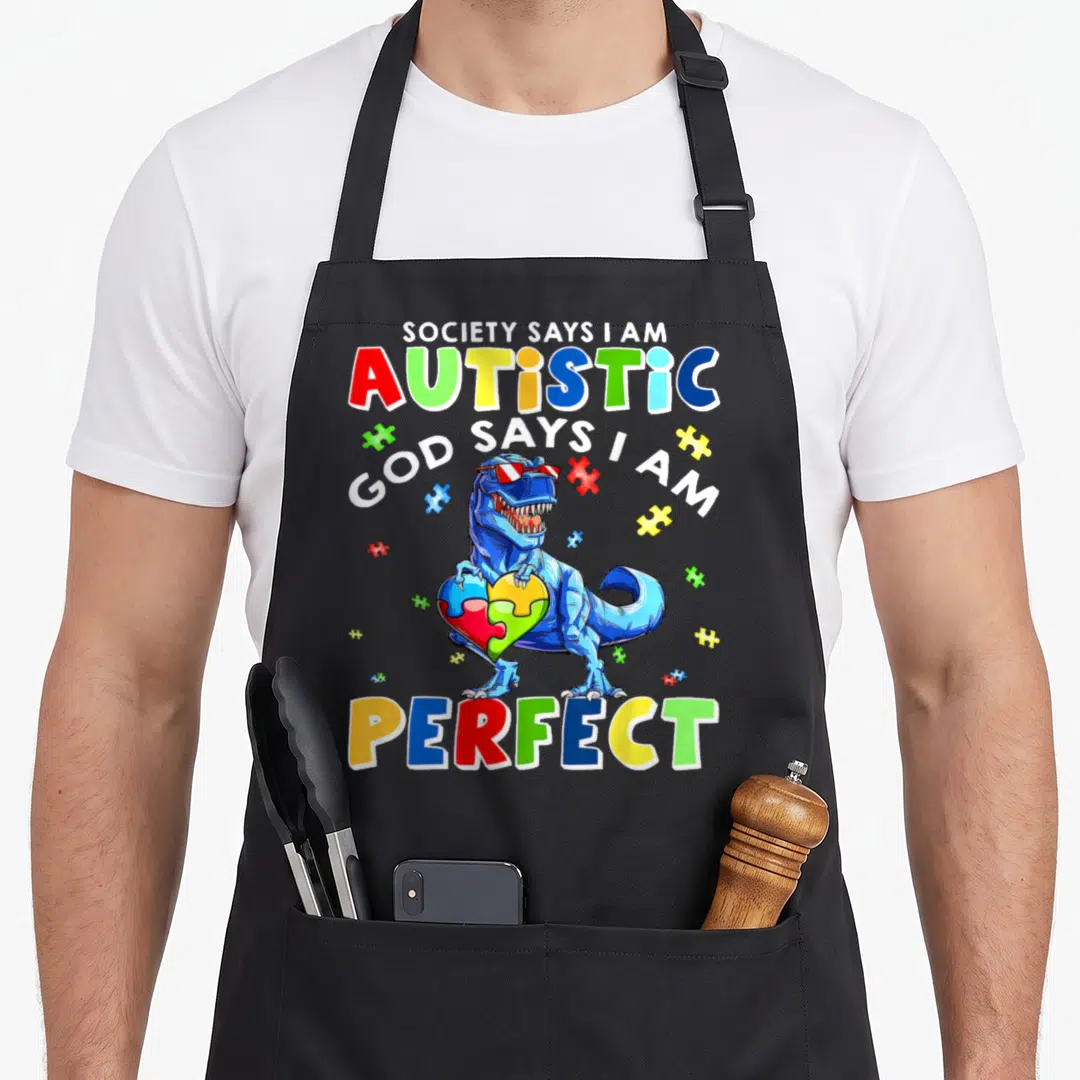 Society tea quotes apron - Blue T-Rex holding puzzle heart with text Society Says I Am Autistic God Says I Am Perfect — shown on a black cotton canvas apron against a white background