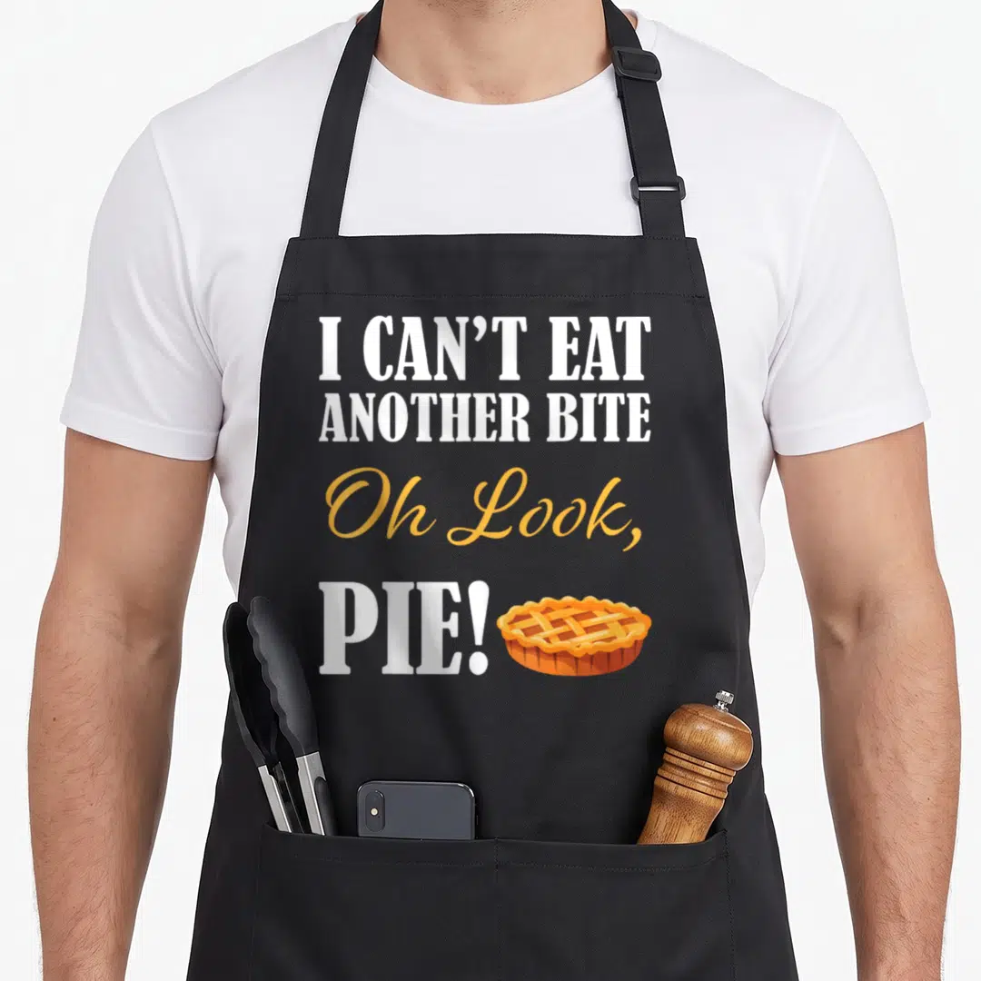 Friendsgiving thanksgiving aprons - I CAN'T EAT ANOTHER BITE Oh Look PIE! text with orange lattice pie drawing — shown on a black cotton canvas apron against a white background