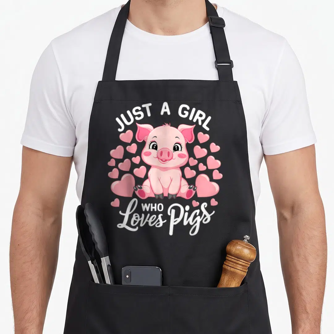 Funny animal i love my pig apron - Cartoon pink pig sitting among hearts with white text Just A Girl Who Loves Pigs — shown on a black cotton canvas apron against a white background