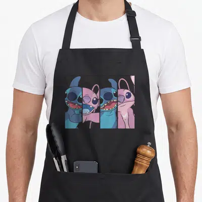 Boxed face apron - Four vertical panels showing blue and pink cartoon characters with large eyes and teeth — shown on a black cotton canvas apron against a white background