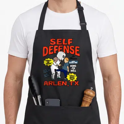 Bobby cartoon apron - Bobby Hill kicking figure with Self Defense for Women text and Arlen TX at bottom — shown on a black cotton canvas apron against a white background